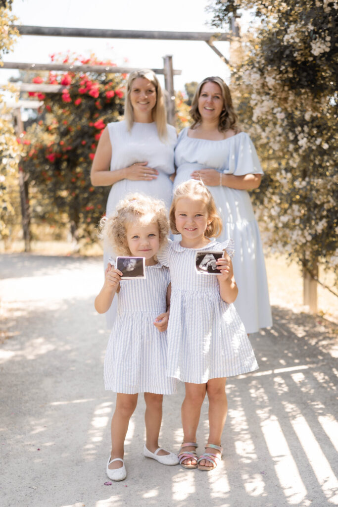 Babybauch-shooting-niederoesterreich-grosse-schwestern-mit-ultraschall-bild-vorne-muetter-im-hintergrund-hand-am-bauch